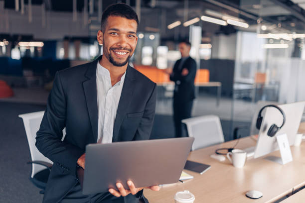 Portrait of african businessman with laptop looking in camera and smiling on colleague background